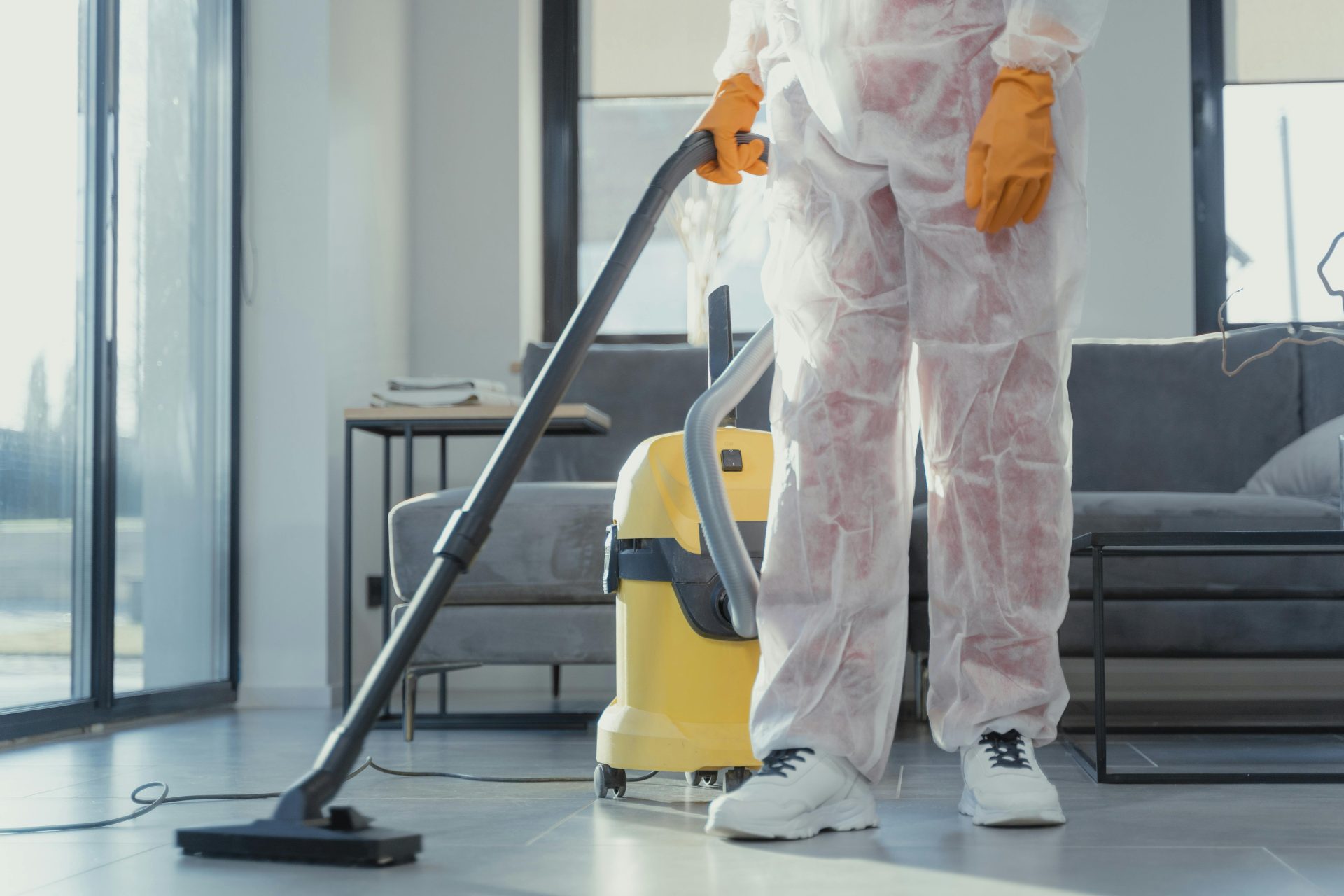 Une femme de ménage en combinaison et gants de protection passe l'aspirateur dans un salon moderne.